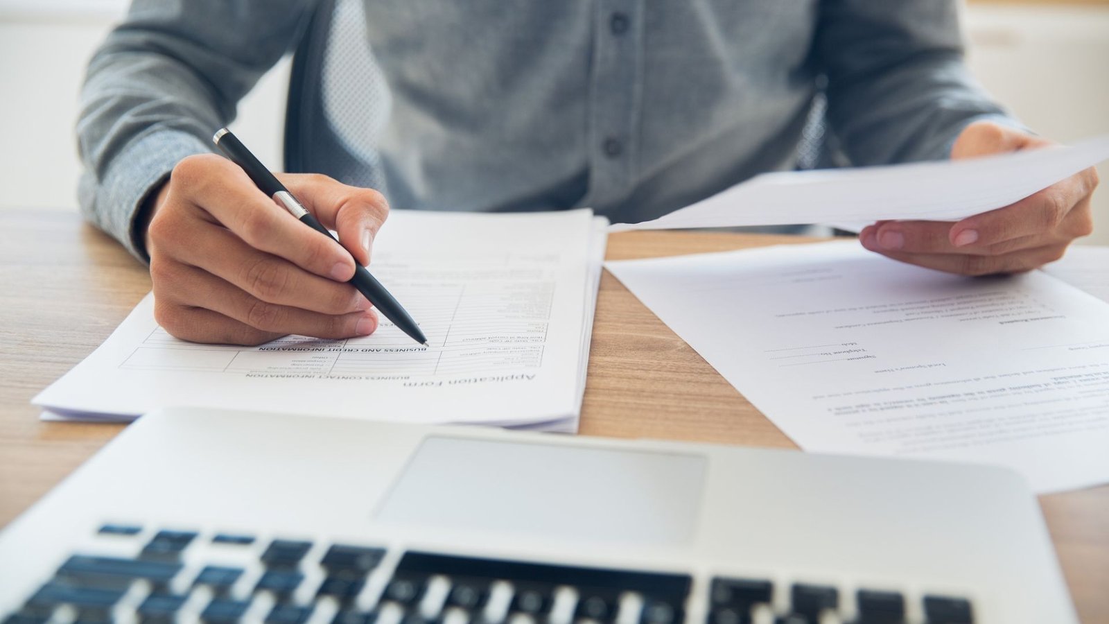 Person reviewing and signing paperwork at a desk next to a laptop